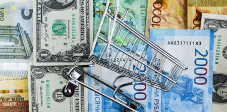 Russian rubles, American dollars, euros, Kazakhstani tenge under an empty grocery cart. International financial crisis. Selective focus.の写真素材