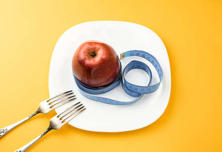 A red apple, wrapped in a centimeter, on a white plate, on a orange background, next to two table forks. The concept of healthy food, fitness, diet, weight loss. View from above. Selective focus.の写真素材
