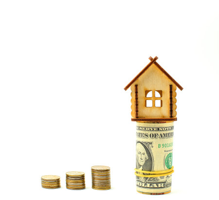 Miniature wooden house stands on top of rolled USA banknotes, metal coins isolated on a white...
