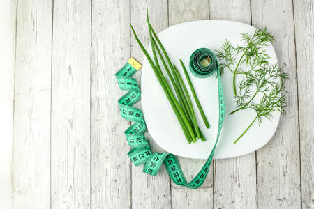 Green onions, dill and tape measure on a white plate, light wooden background. Concept of healthy food, fitness, diet, weight loss. Top view. Flat lay.の写真素材