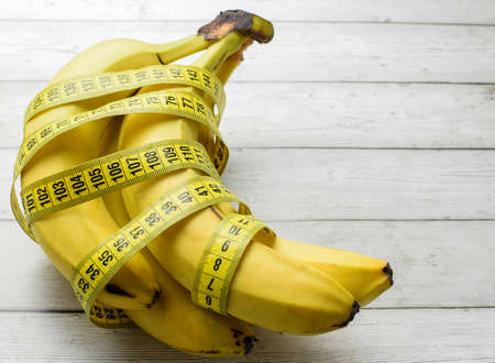 Ripe yellow bananas, wrapped with a centimeter tape, on a light wooden background. The concept of healthy food, fitness, diet, weight loss. Selective focus.の写真素材