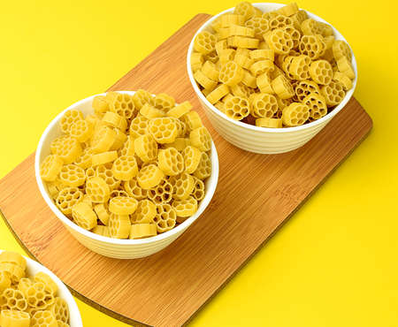 Raw pasta in a white bowl on a wooden cutting board, bright yellow background. Copy space for your...
