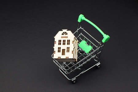 Wooden house in a supermarket trolley on a black background. Real estate buying and selling concept. Renting out the property. Copy space.の写真素材