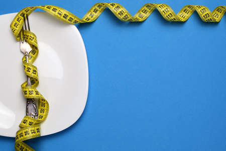 Bright measuring tape on a white plate, a fork on a colored background. The concept of losing weight, diet, proper nutrition, healthy lifestyle. Top view, flat lay. Copy space for text.の写真素材