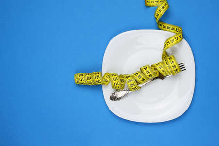 Fork is wrapped in a centimeter tape, lies on a white plate, on a blue background. Concept of weight loss, diet, proper nutrition, healthy lifestyle. Top view. flat lay. Selective focus.の写真素材