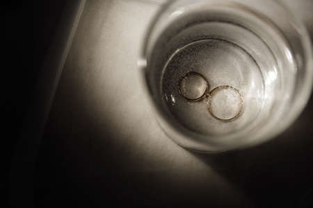 Two gold wedding rings close-up at the bottom of a glass of sparkling water. White veil on the background.の写真素材