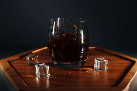There are ice cubes and an alcoholic cocktail on a wooden tray. There are interesting waves and splashes in the glass. Fluid in motion.の写真素材