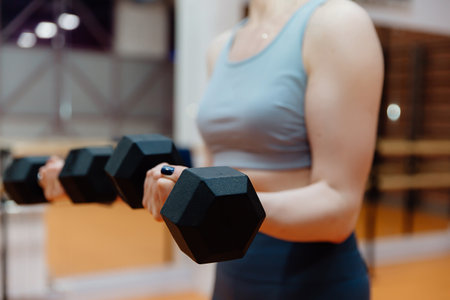 A sporty girl in a gray top with pumped-up arms performs an exercise with dumbbells. Sports in the fitness club.の写真素材