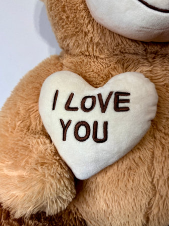 A white heart-shaped plush or soft material with 'I LOVE YOU' text embroidered in dark letters, placed against a light brown teddy bear or plush toy backgroundの写真素材