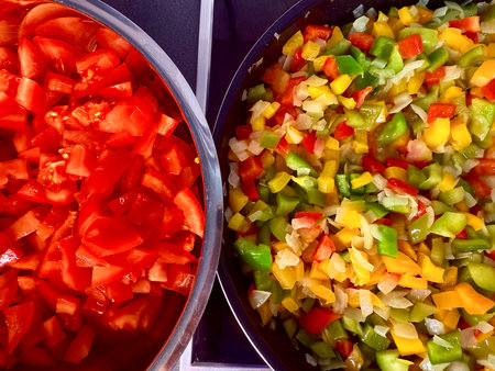 Chopped vegetables in a bowl, fresh ingredients for cooking preparation: tomato, bell pepper, and onion. Kitchen cloth, cutting board, and knife on marble countertop.の写真素材