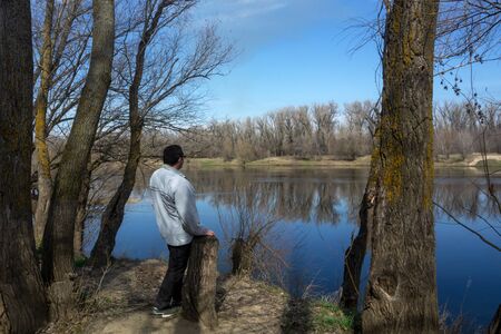 A middle-aged man stands with his back and looks at the spring river in the forest. Bright sky and...