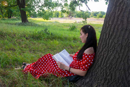 Young woman is resting on a summer day in the park. A brunette with a book on the grass near a tree sits with her back to the trunk. Summer mood, harmony, outdoor recreation.の写真素材