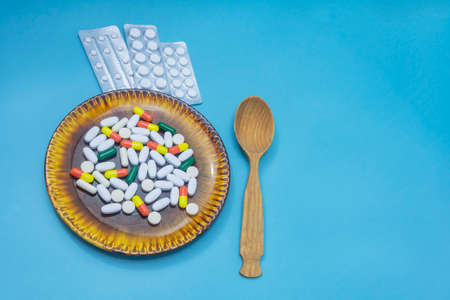 Many different pills and capsules of medicines on dark plate. Wooden spoon on light blue background. The concept of overuse medicines, antibiotics, vitamins, aspirin.の写真素材