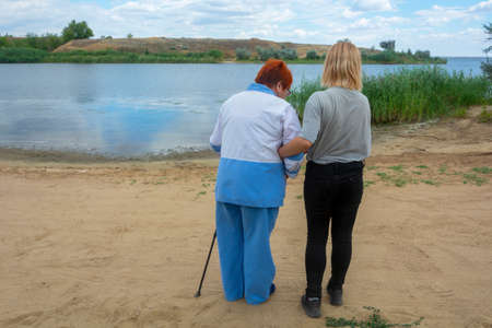 Young woman helps her older grandmother to walk. Granddaughter supports grandmother outdoors....