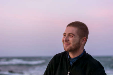 Smiling young Caucasian man with beard on background of the sea and waves with white foam. Selective...