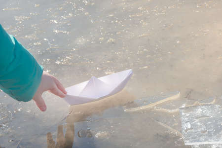 Child's hand launches white paper boat into puddle of ice. Early spring mood concept, freedom.の写真素材