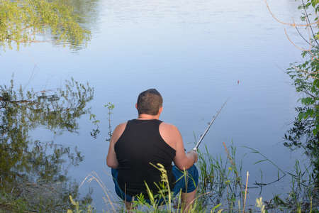 Male fisherman sits with fishing rod on bank of river and catches fish. View from back.