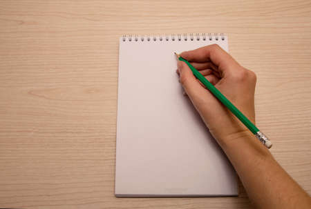 Woman's right hand writes in green pencil in white notebook on a wooden backgroundの写真素材