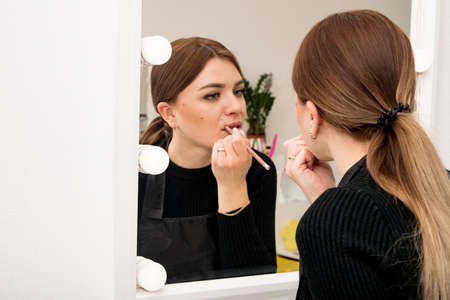 Young blonde girl in black tints lips with a pencil in the mirror