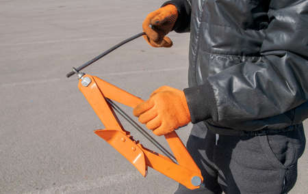 Metal orange small foot jack with handle for car in hands of man wearing orange gloves.