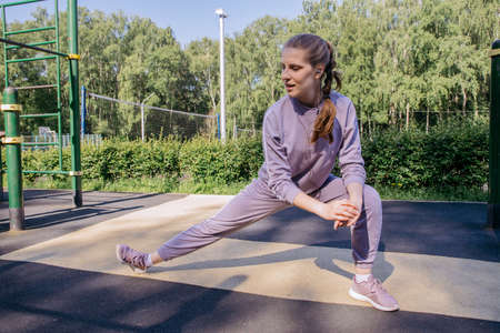 A girl in headphones and a sports uniform on the playground performs a leg stretch