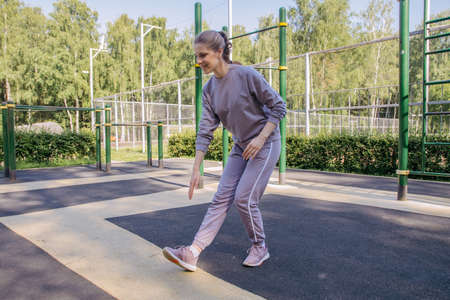 A girl in headphones and a sports uniform on the playground performs a leg stretch exercise