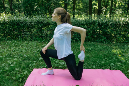 A girl in a white t-shirt and black leggings on a pink mat on a summer green lawn and doing yoga