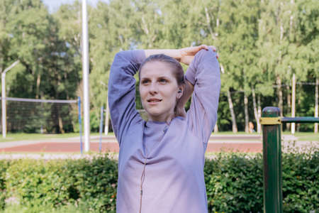 Girl in headphones and sports uniform on the playground stretches the biceps of the arms