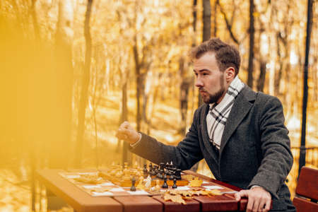 One European man sits at a chess table on a playground in an autumn park with fallen yellow leaves and plays with himselfの写真素材