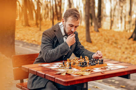 One European man sits at a chess table on a playground in an autumn park with fallen yellow leaves and plays with himselfの写真素材