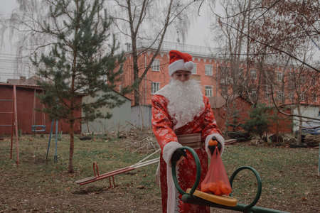 a sad man dressed as Santa Claus with tangerines in a bag stands on the playground. The...