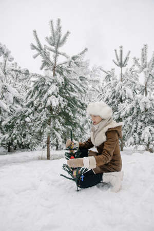A girl in a sheepskin coat, mittens, a white hat with earflaps and boots sits around snow-covered pine trees in winter and dresses up a small artificial Christmas tree with New Year's decorationsの写真素材