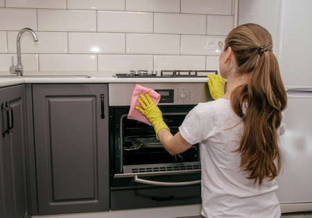 Young brunette girl in rubber yellow gloves rubs the stove oven with a rag in a gray and white kitchenの写真素材