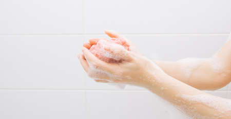 A girl in a white bathroom in foam rubs her hands with a pink washclothの写真素材