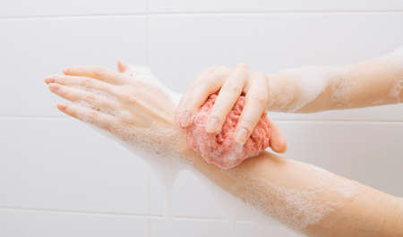 A girl in a white bathroom in foam rubs her hands with a pink washclothの写真素材