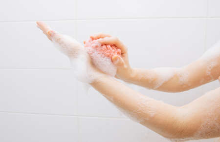 A girl in a white bathroom in foam rubs her hands with a pink washcloth