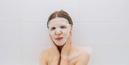 Girl in a white bathroom with foam makes a moisturizing sheet mask for the faceの写真素材