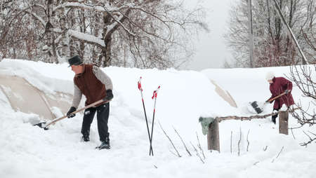 01.29.2022. Russia, Tula, Skuratovo village. An elderly man and a woman remove snow from the roof of the greenhouse with a shovel in the courtyard of the house in winterの写真素材