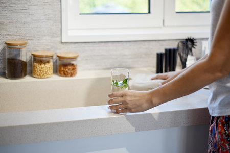 A woman holding a glass of water near the sink in the kitchenの写真素材
