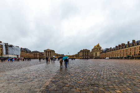 Versailles, France - May 28, 2024: Visitors stroll through the expansive cobblestone courtyard of the Palace of Versailles, France, under umbrellas on a rainy day, taking in the grandeur of this historic siteのeditorial素材