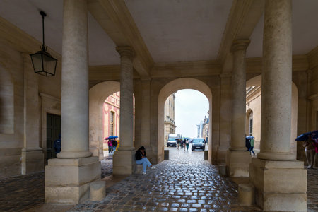Versailles, France - May 28, 2024: Visitors at the Palace of Versailles seek shelter from the rain under the grand arched passageway, with cobblestones glistening in the wet weatherのeditorial素材