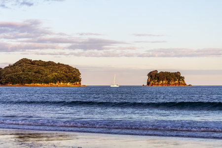 A sailboat gently glides between two lush, forested islands as the sun sets over the tranquil waters of Whangamata, New Zealandの写真素材