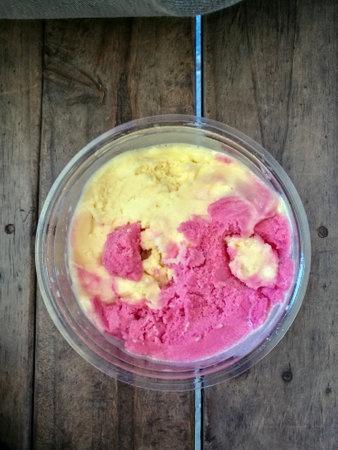 A vibrant bowl of pink and yellow ice cream served in a clear container on a rustic wooden surfaceの写真素材
