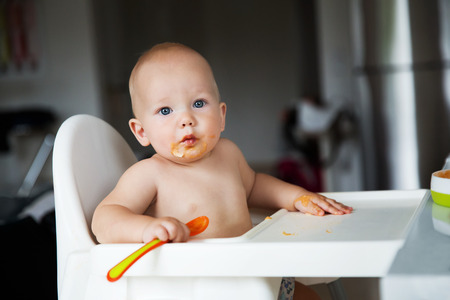 Feeding. Adorable baby child eating with a spoon in high chair. Baby's first solid foodの写真素材
