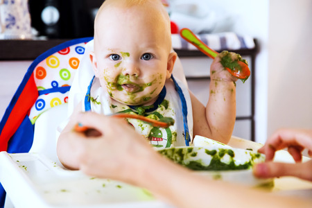 Feeding. Adorable baby child eating with a spoon in high chair. Baby's first solid foodの写真素材