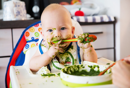 Feeding. Adorable baby child eating with a spoon in high chair. Baby's first solid foodの写真素材