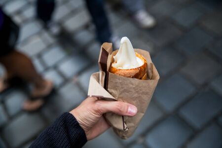 Tourist holds in hand Trdlo or Trdelnik with ice-cream on the background of city tourist streets of Czech (Prague). Fresh Appetizing Trdlo or Trdelnik - Traditional National Czech Sweet Pastry Dough.の写真素材