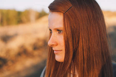 Portrait of Beauty Teenage Model Girl with Red Hair on the Background of Nature on the Field in Sun Light. Face of Young Woman with Freckles. Autumn. Glow Sun, Sunshine. Backlit. Warm Color Tonesの写真素材