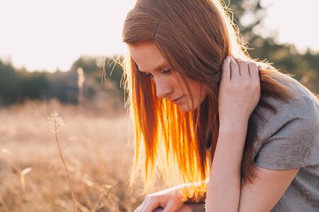 Portrait of Beauty Teenage Model Girl with Red Hair on the Background of Nature on the Field in Sun Light. Face of Young Woman with Freckles. Autumn. Glow Sun, Sunshine. Backlit. Warm Color Tonesの写真素材