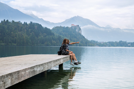 Young couple of tourists in love sitting on a wooden pier on the background with Bled Castle and Church on the Island on the Lake Bled, Slovenia. Autumn time in Europe.の写真素材